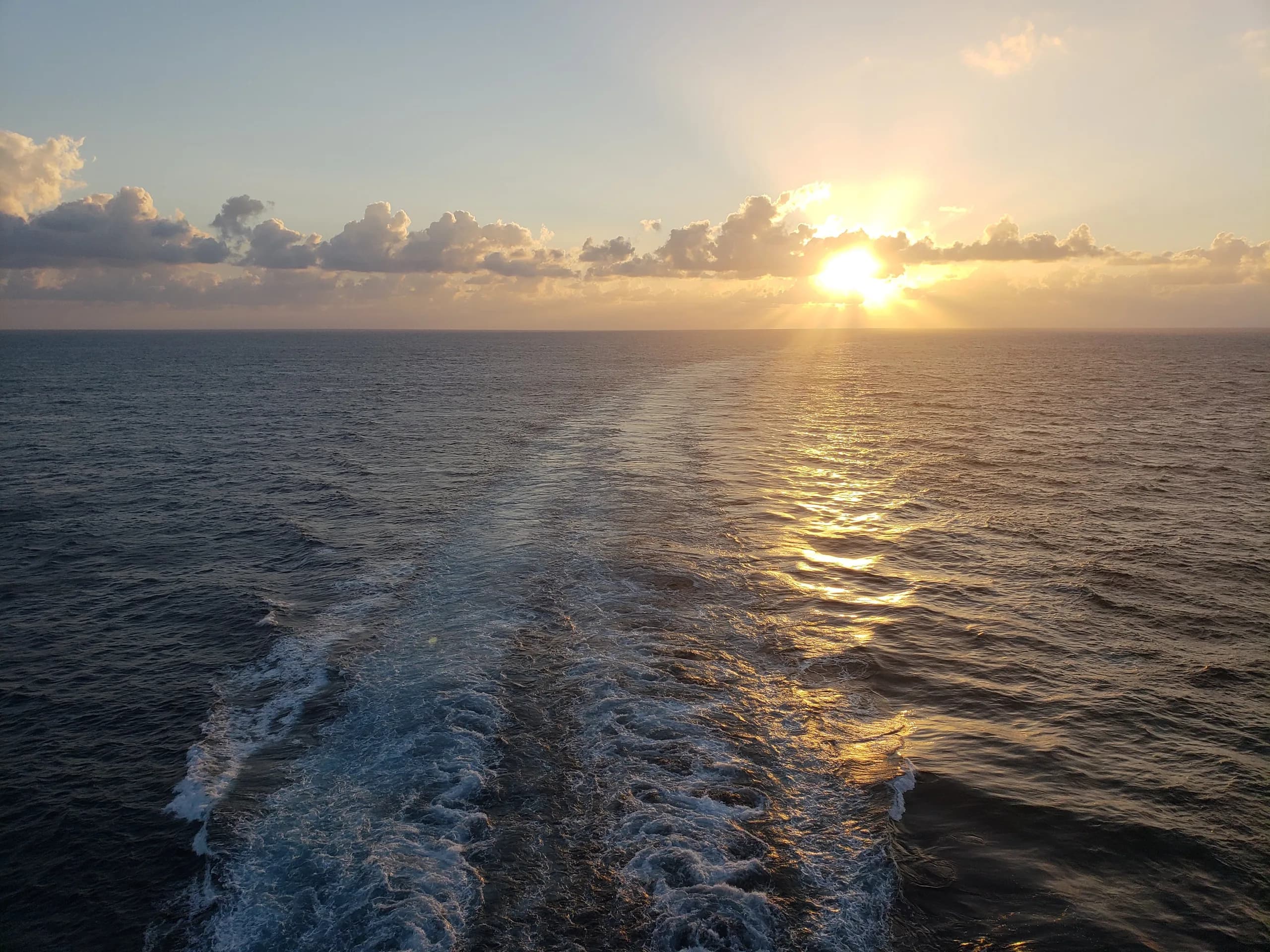 The sun low over the open sea, seen from the back of a cruise ship, with the ship's wake trailing toward it