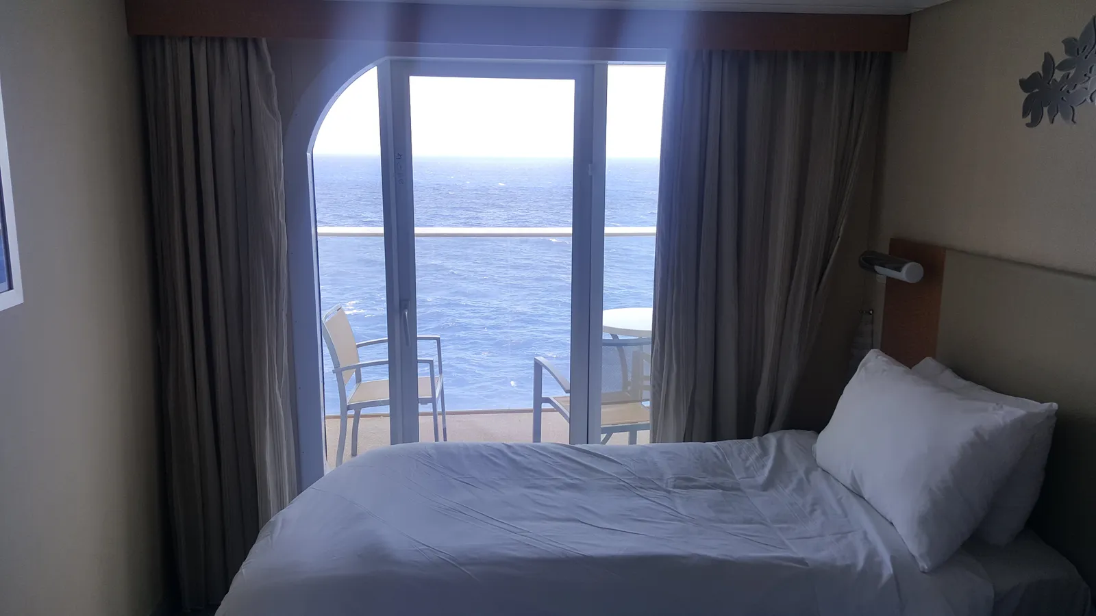 A balcony stateroom on Oasis of the Seas: the bed faces a sliding glass door, with two chairs and a small table on the balcony and open sea beyond